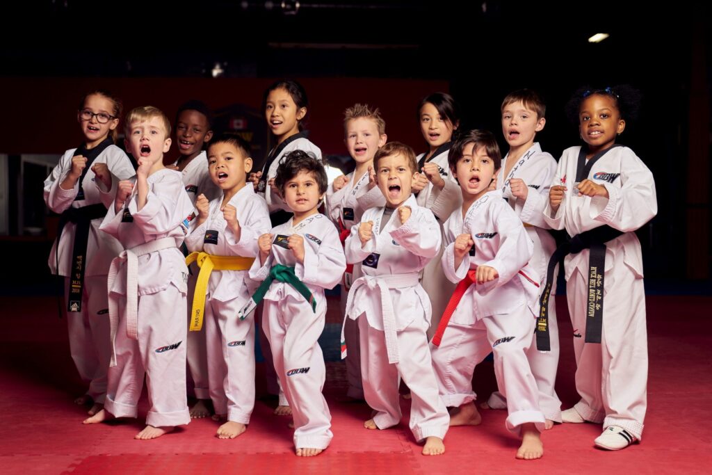 A diverse group of young martial arts students wearing white uniforms and various colored belts practicing their kihap shouts during a kids' karate class.
