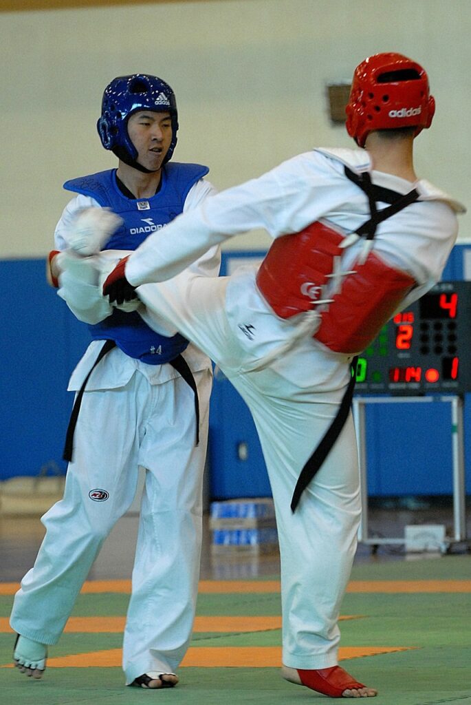 Two Taekwondo athletes in white doboks and full protective gear engaged in a strategic point gap match featuring a high-scoring turning kick attempt.