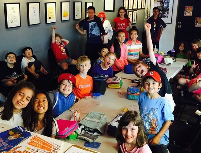 A group of happy children sitting at long tables during an indoor youth program focused on healthy socialization.