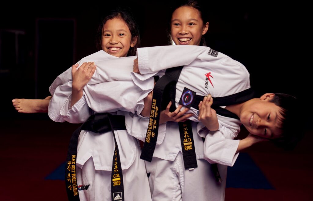 Two young girls laughing while lifting their friend to demonstrate teamwork and friendship in their youth karate class.
