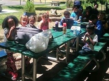 A group of children enjoying a healthy lunch outdoors during an active youth camp to foster social development.
