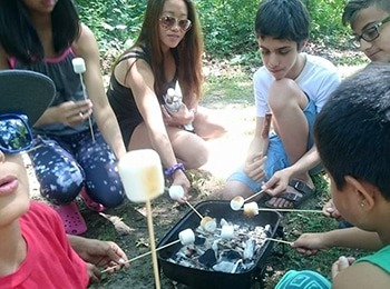 A group of children and a counselor roasting marshmallows over a small grill during a youth summer program to foster teamwork and friendship.